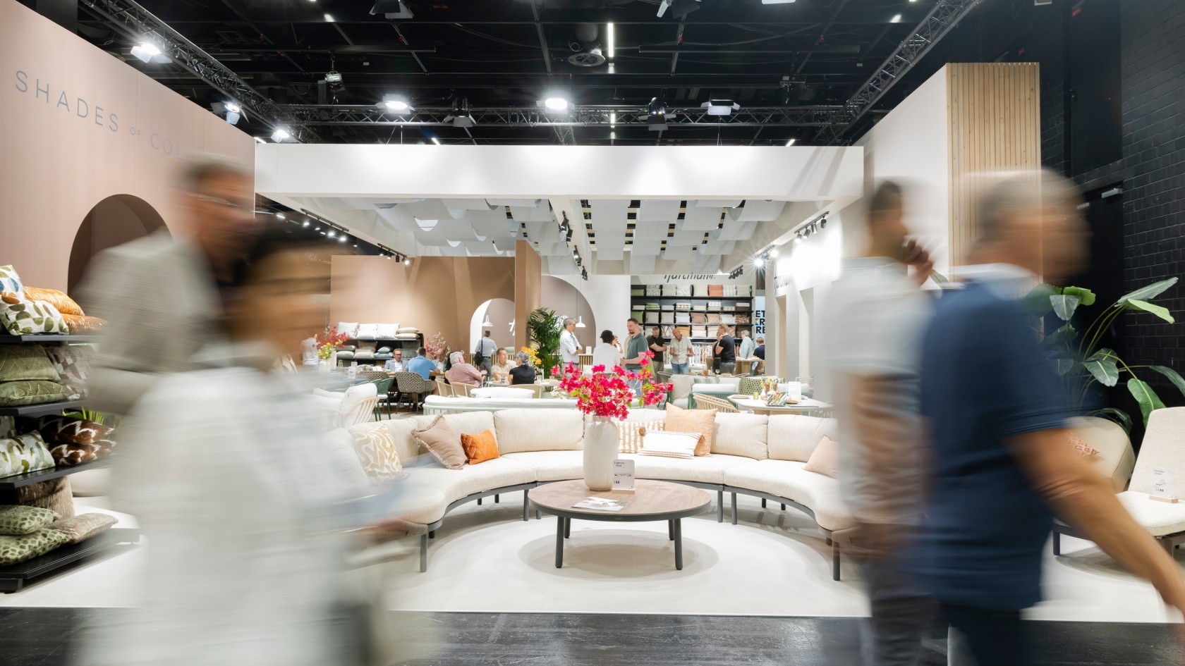 A lively trade show area featuring a curved couch, decorative flowers, and exhibits, with attendees moving through the space.