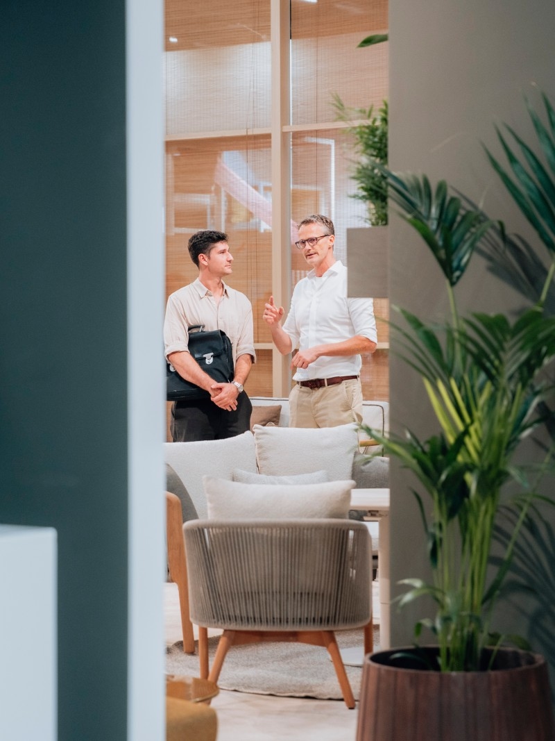 Two men are engaged in conversation in a modern, stylish room with plants and comfortable seating.