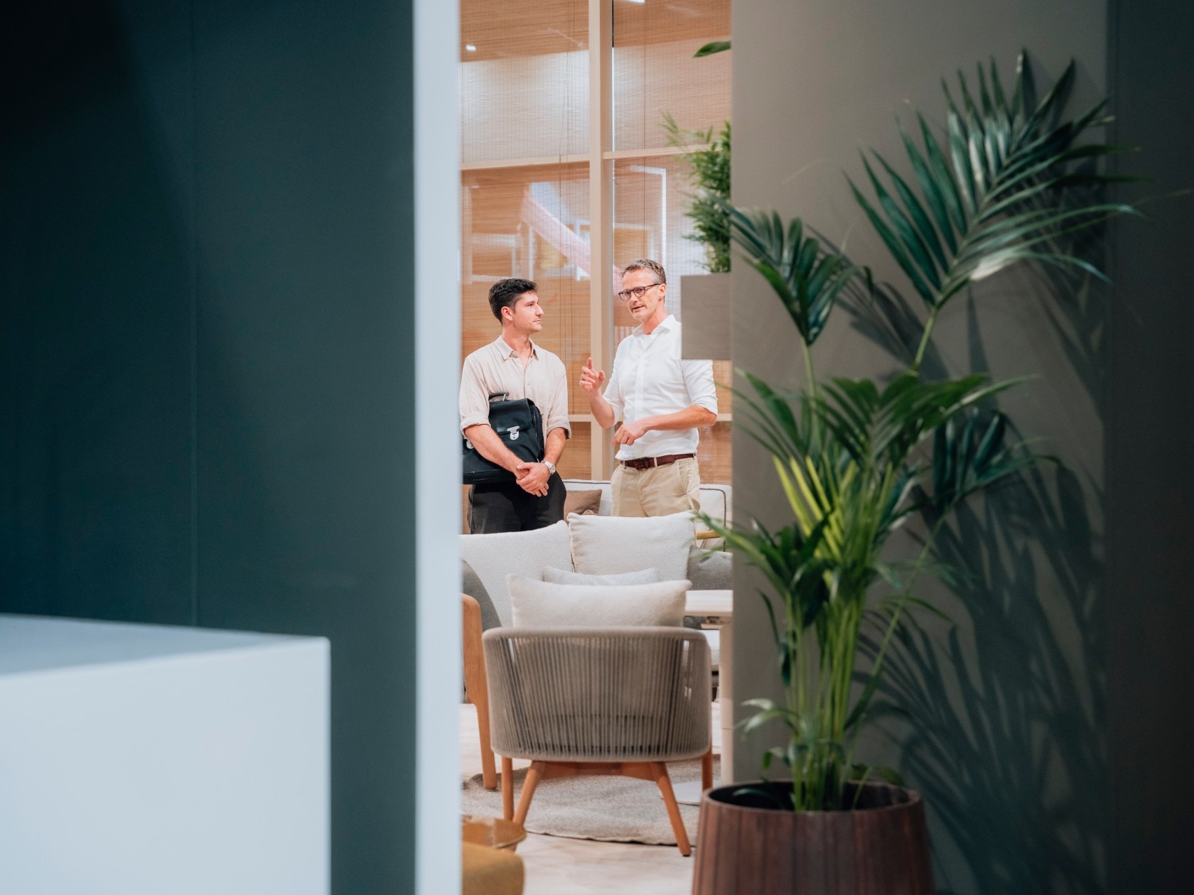 Two men are engaged in conversation in a modern, stylish room with plants and comfortable seating.