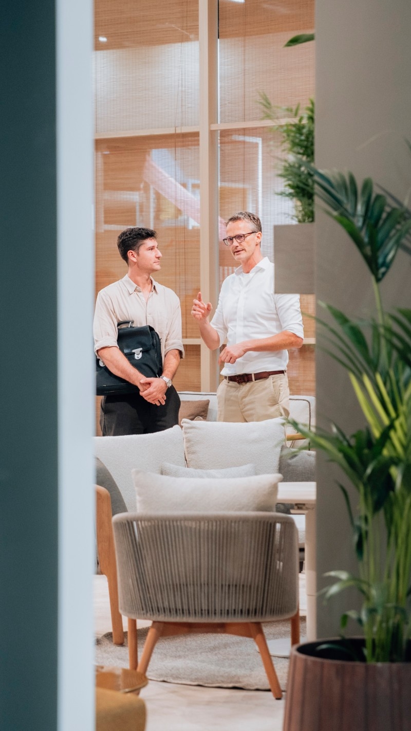 Two men are engaged in conversation in a modern, stylish room with plants and comfortable seating.