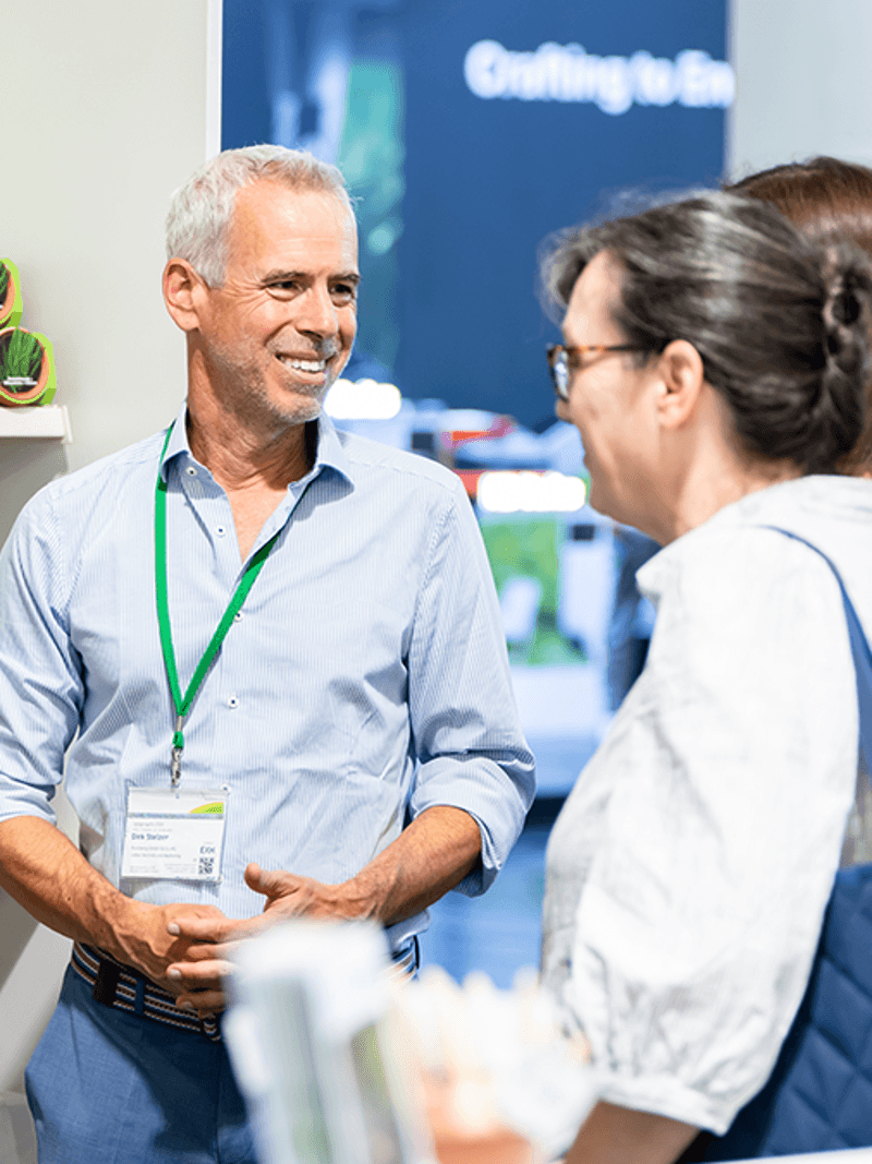 A man in a blue shirt converses with two women at a booth showcasing eco-friendly products, with packaging displayed behind them.