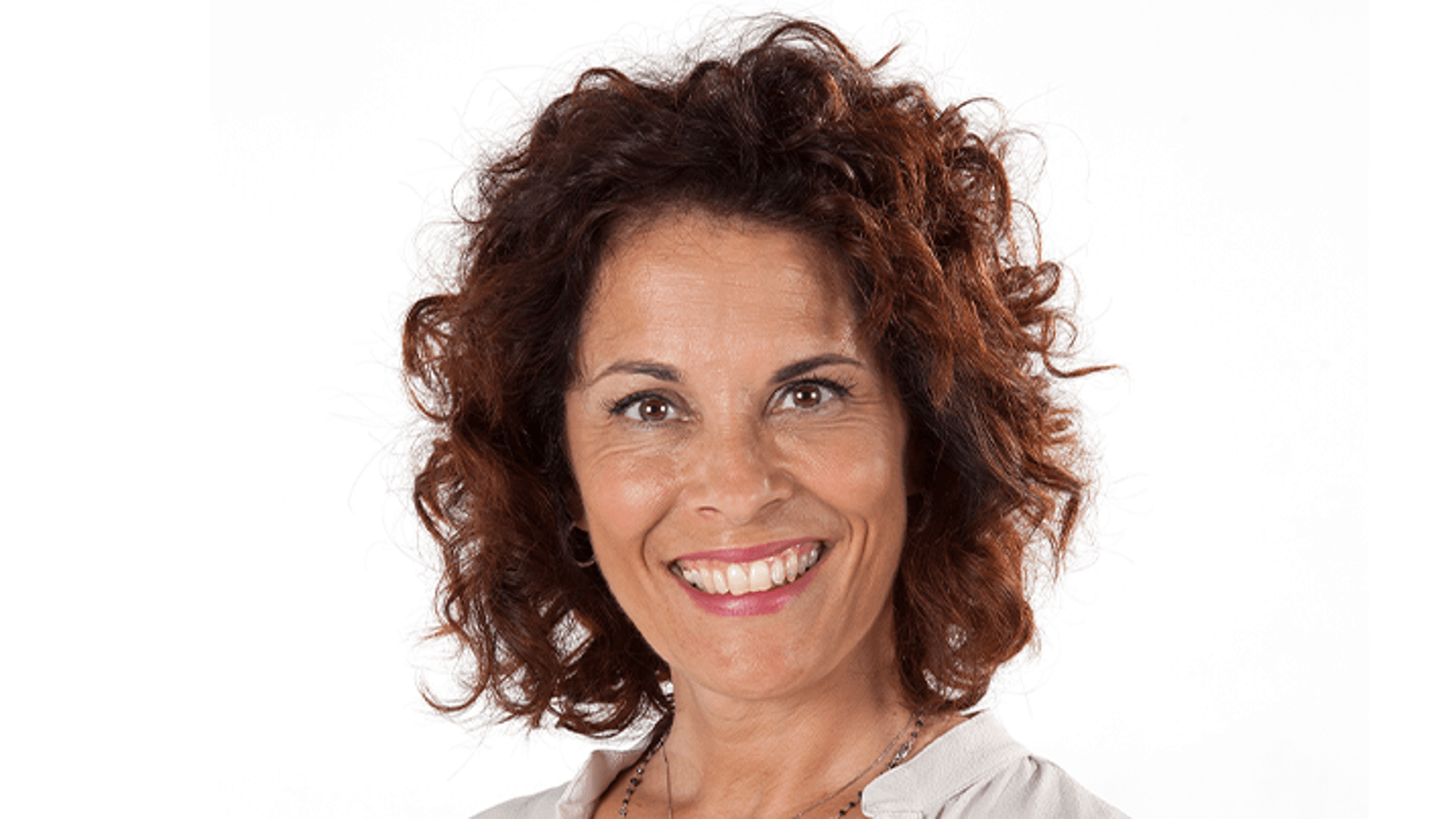 A smiling woman with curly hair stands in front of a white background, wearing a light-colored blouse and a necklace.