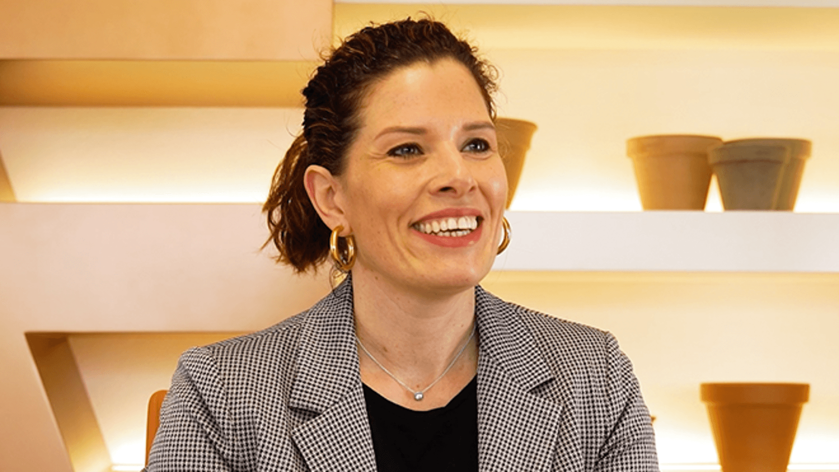 A woman with short, curly hair smiles while sitting at a table, wearing a patterned blazer, with plant pots in the background.