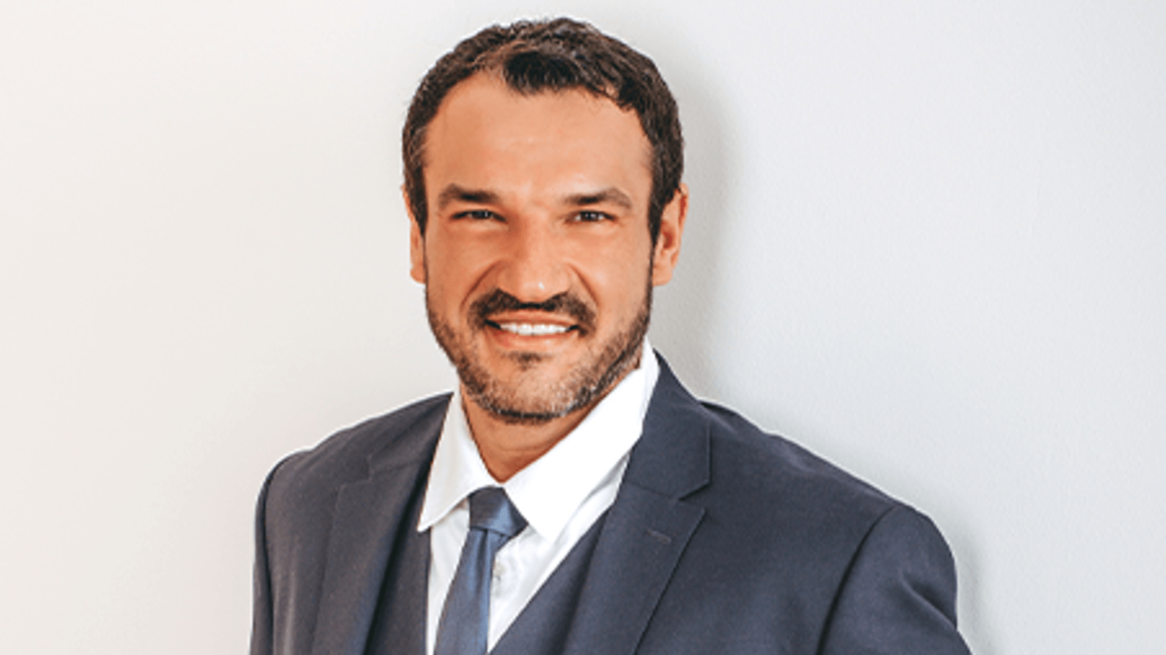 A man in a formal dark suit smiles confidently while standing against a light backdrop, hands in pockets.