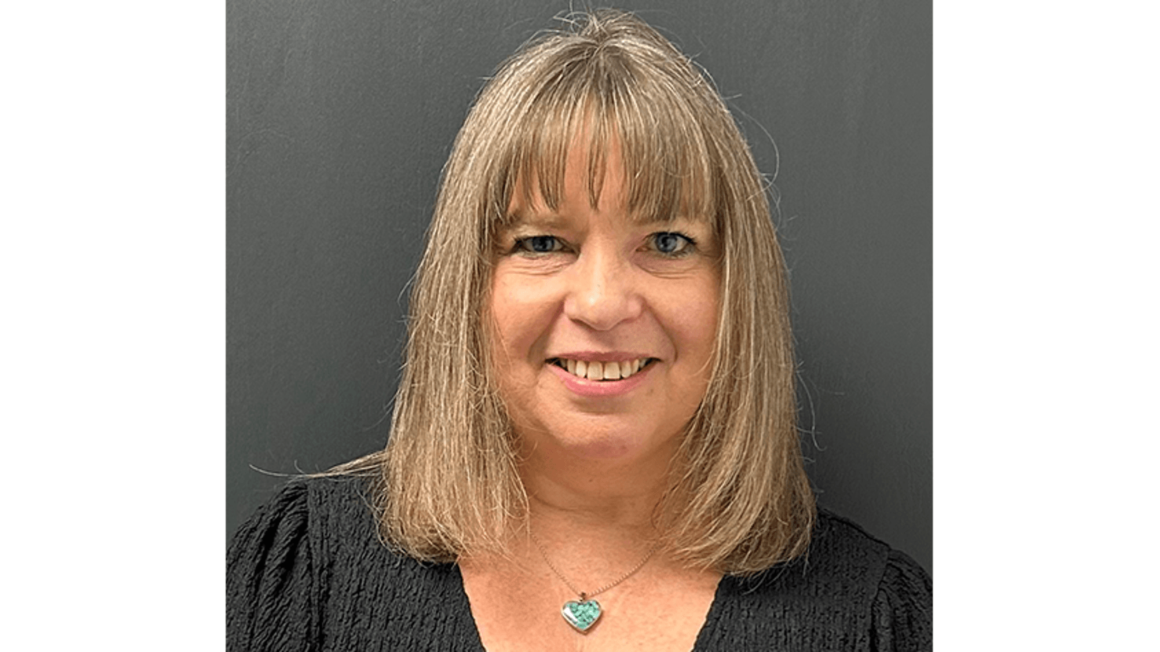 A smiling woman with shoulder-length straight hair, wearing a black top and a turquoise necklace, stands against a dark gray background.