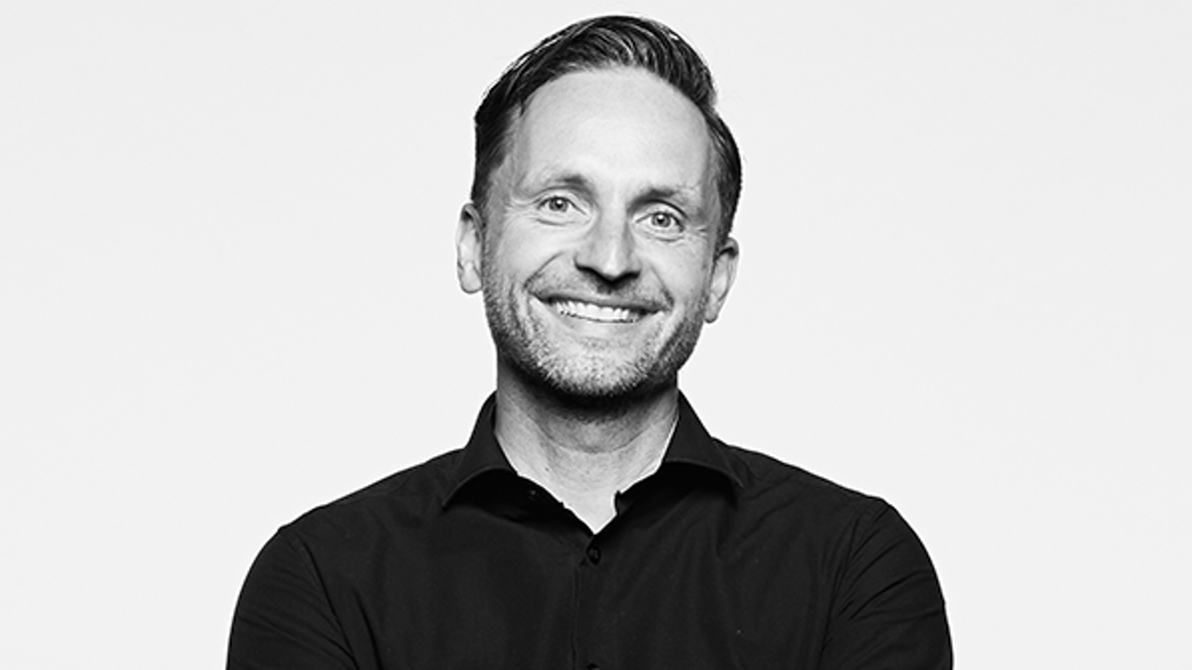A smiling man in a black shirt stands with arms crossed against a plain white background.