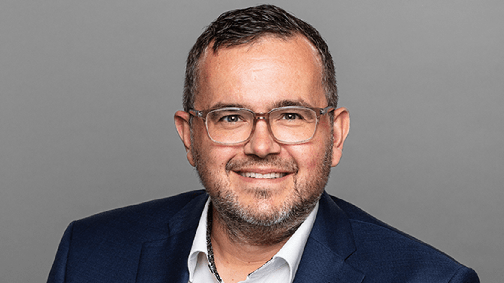 A smiling man with glasses, wearing a blue suit over a white shirt, poses against a gray background. His expression is friendly and approachable.