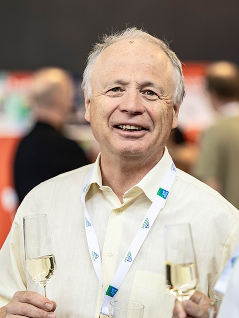 Zwei Männer führen ein Gespräch, jeder hält ein Glas Champagner, lächeln vor einem lebhaften Hintergrund mit Veranstaltungsausstellungen.