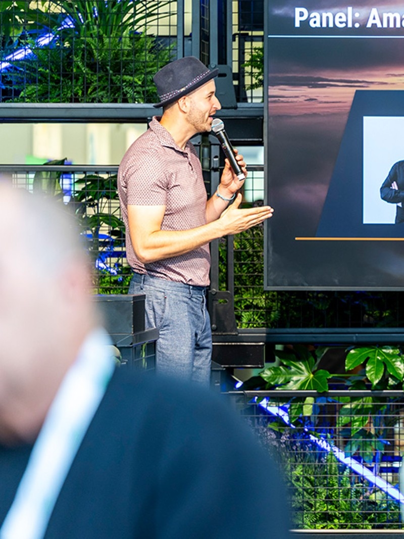 Two presenters are on stage discussing a panel titled "Amazon im Wandel," with a display featuring William Cobussen's contact details.