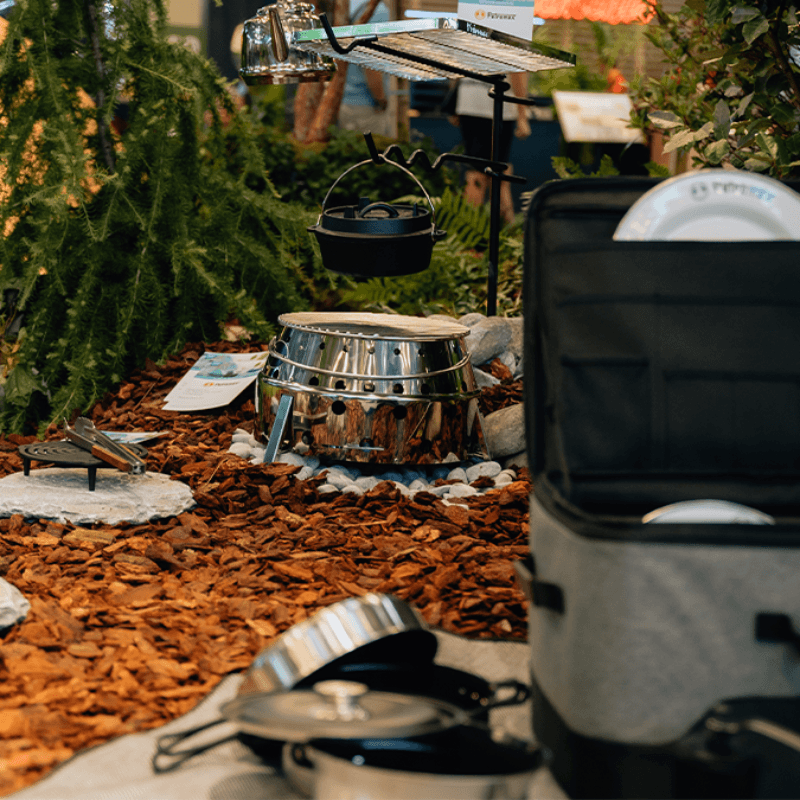 Eine Ausstellung des Campingplatzes zeigt Kochgeschirr, Steinwege und einen weichen Kühler, der von dekorativer Mulch und Grün umgeben ist.