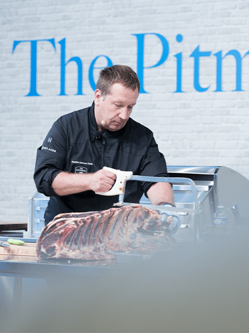 Ein Koch in schwarzer Uniform verwendet eine Säge, um Fleisch bei einer Kochvorführung zuzubereiten, mit dem Schild "Der Grillmeister" im Hintergrund.