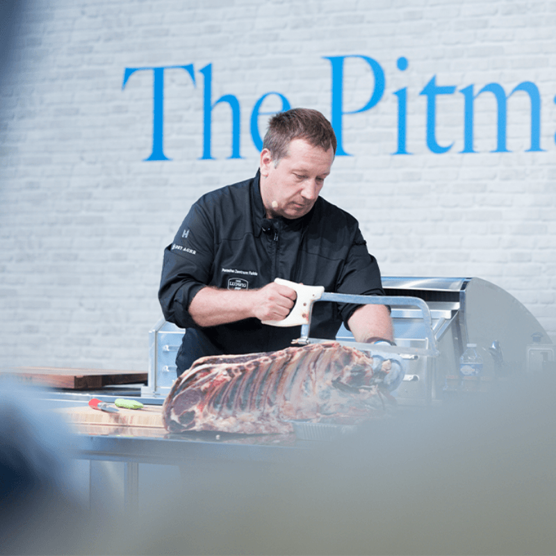Ein Koch in schwarzer Uniform verwendet eine Säge, um Fleisch bei einer Kochvorführung zuzubereiten, mit dem Schild "Der Grillmeister" im Hintergrund.