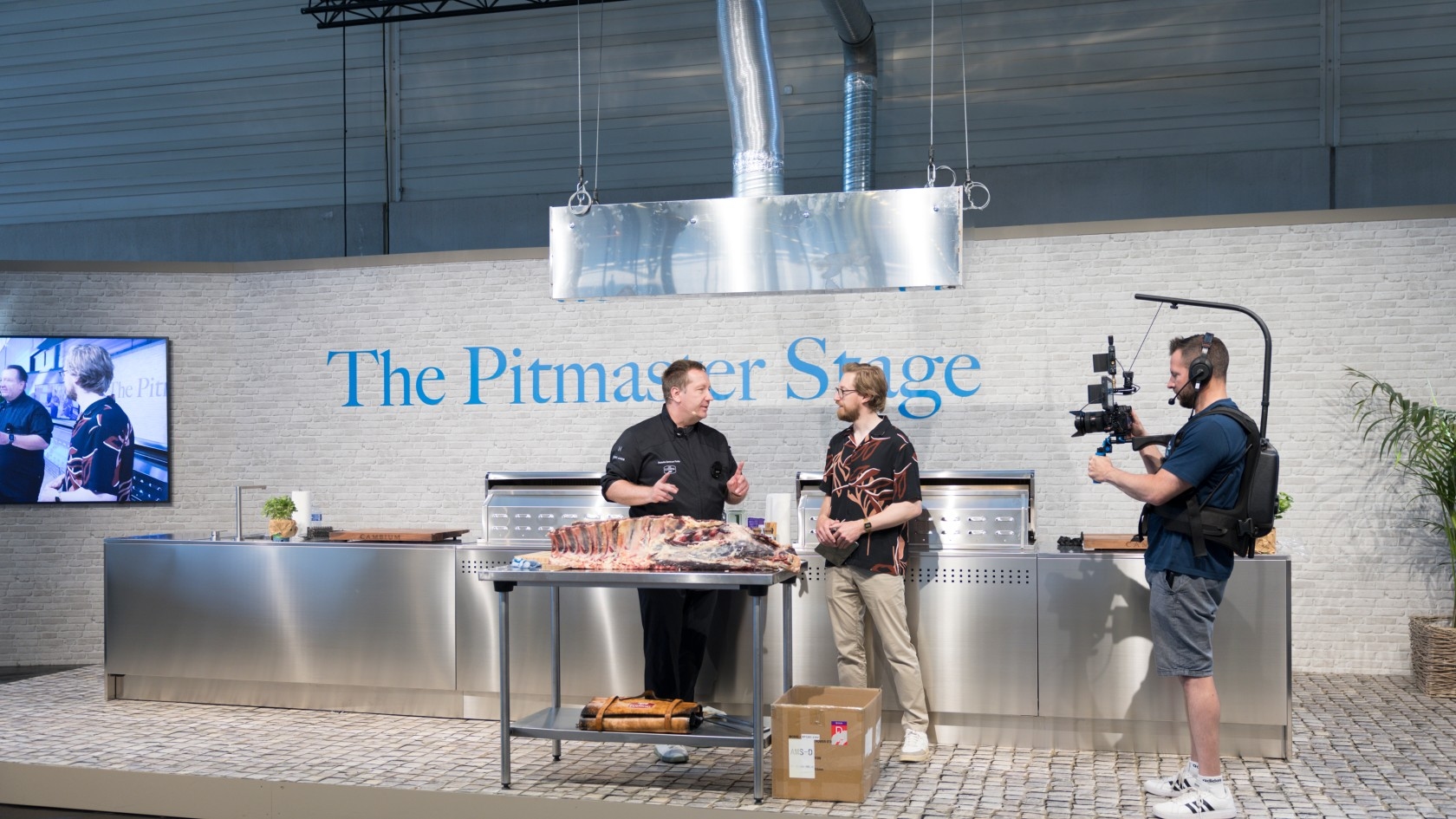 Zwei Männer, einer kocht und der andere führt ein Interview, stehen neben einem Grilldisplay mit der Aufschrift "Die Pitmaster-Bühne", während ein Kameramann sie filmt.