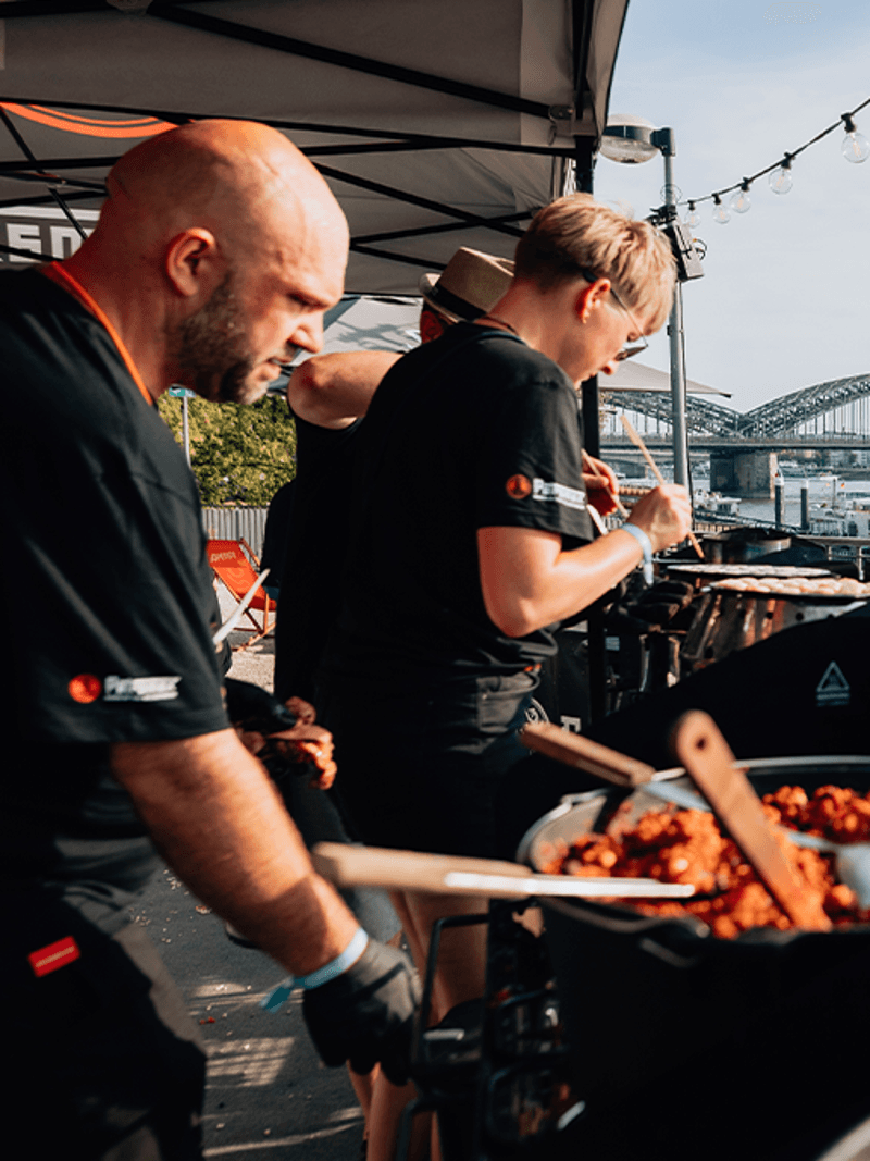Köche bereiten Essen bei einer Outdoor-Veranstaltung am Wasser zu, mit dem Kölner Dom und der Hohenzollernbrücke im Hintergrund.