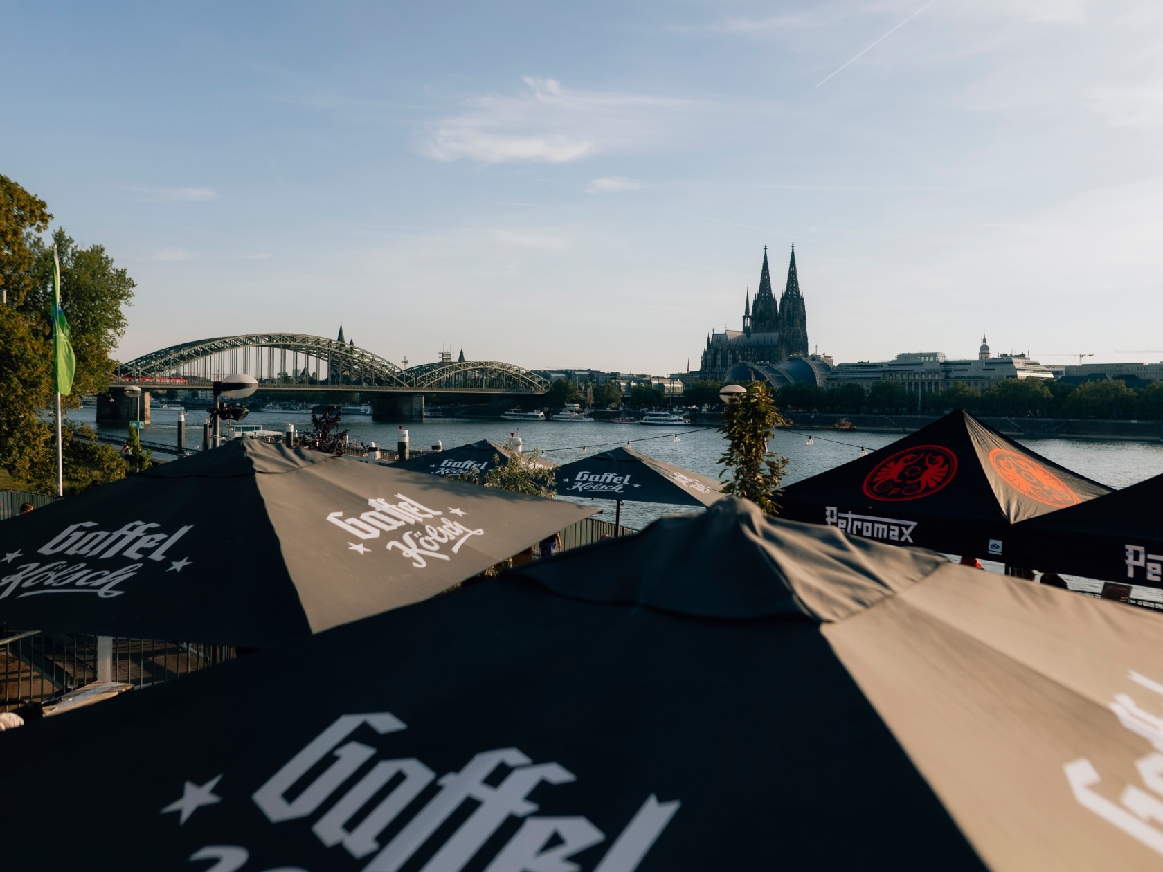 View of Cologne's skyline featuring the cathedral and bridge, with shaded umbrellas in the foreground along the riverbank.