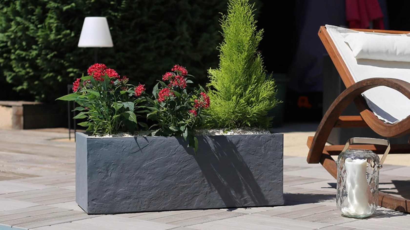 Ein grauer Blumentopf mit roten Blumen und einem kleinen grünen Baum steht neben einem Holzliegestuhl und einer gläsernen Laterne auf einer Terrasse.