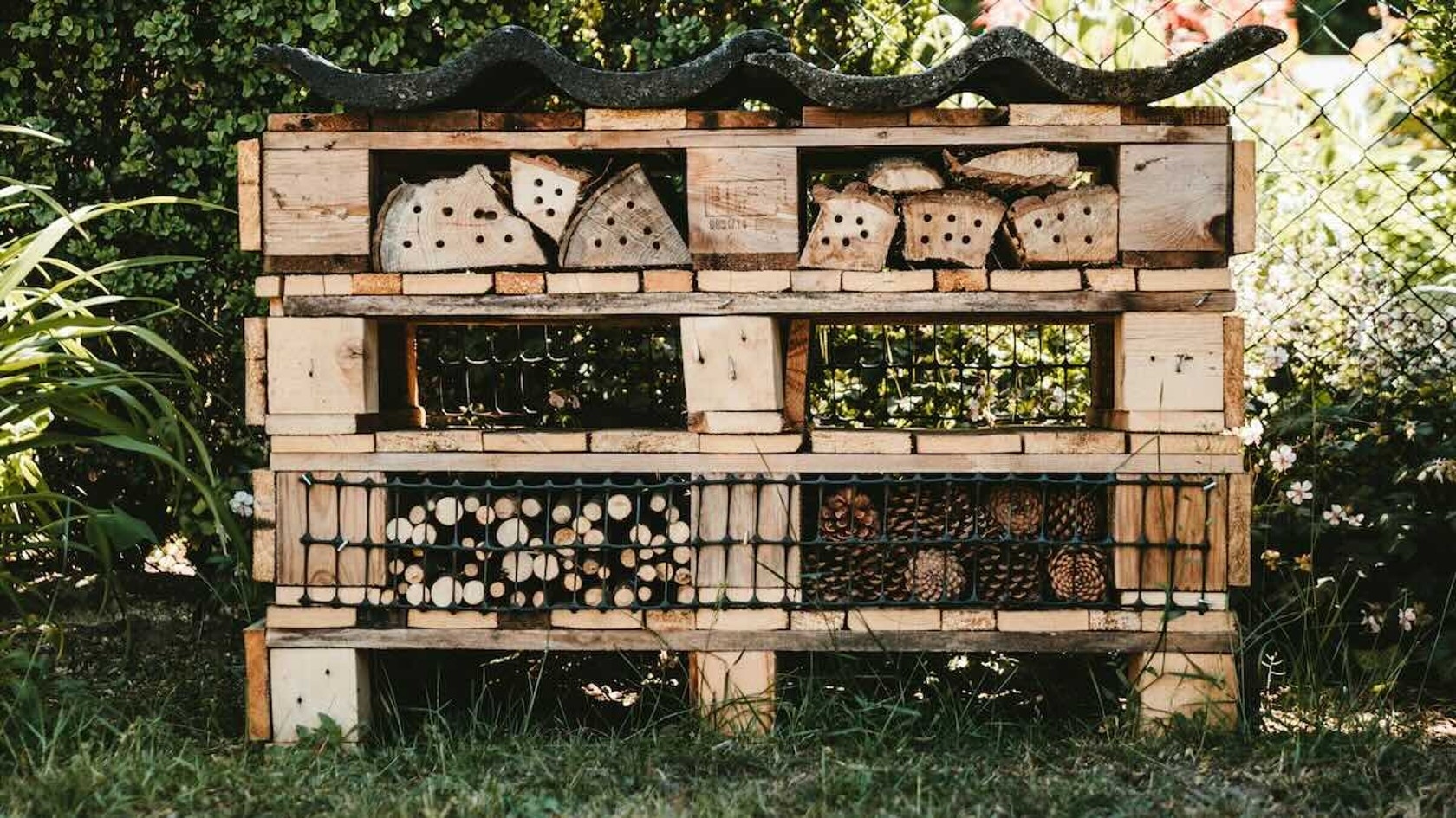 Ein hölzernes Insektenhotel mit verschiedenen natürlichen Materialien, darunter Baumstämme, Tannenzapfen und strukturiertes Holz, in einem Garten aufgestellt.