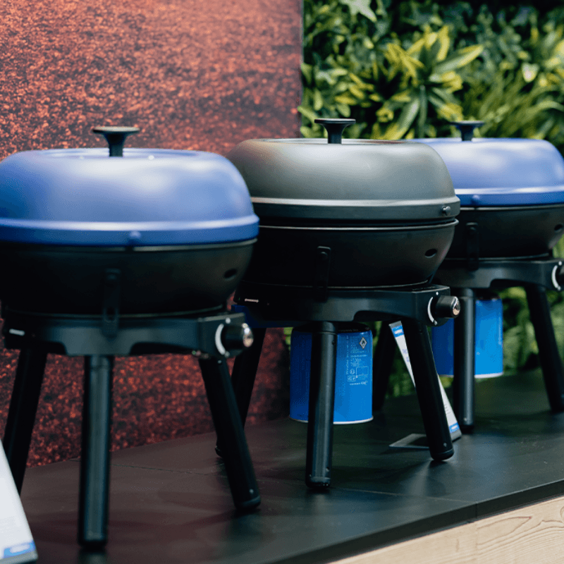 spoga+gafa 2026 Tag 3 06 Three compact outdoor grills in blue and black, displayed on a table, with product information cards in front and a greenery backdrop.