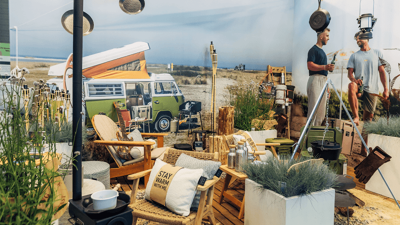 Ein gemütlicher Campingplatz mit Stühlen, Laternen und einem alten Wohnmobil vor einer malerischen Outdoor-Kulisse, die Entspannung und Abenteuer suggeriert.